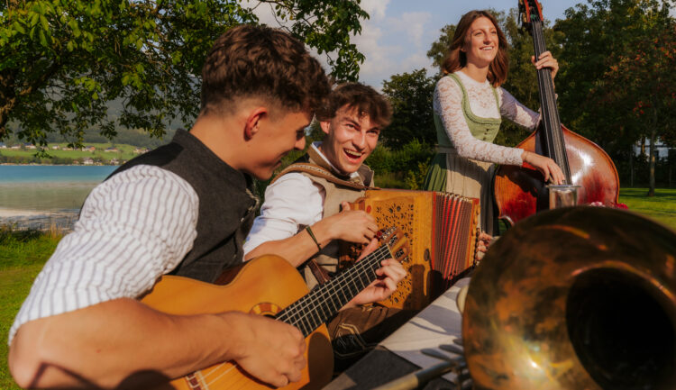 Drei junge Musiker spielen Gitarre, Akkordeon und Kontrabass an einem sonnigen Tag im Freien, lächeln und haben Spaß an der Musik.