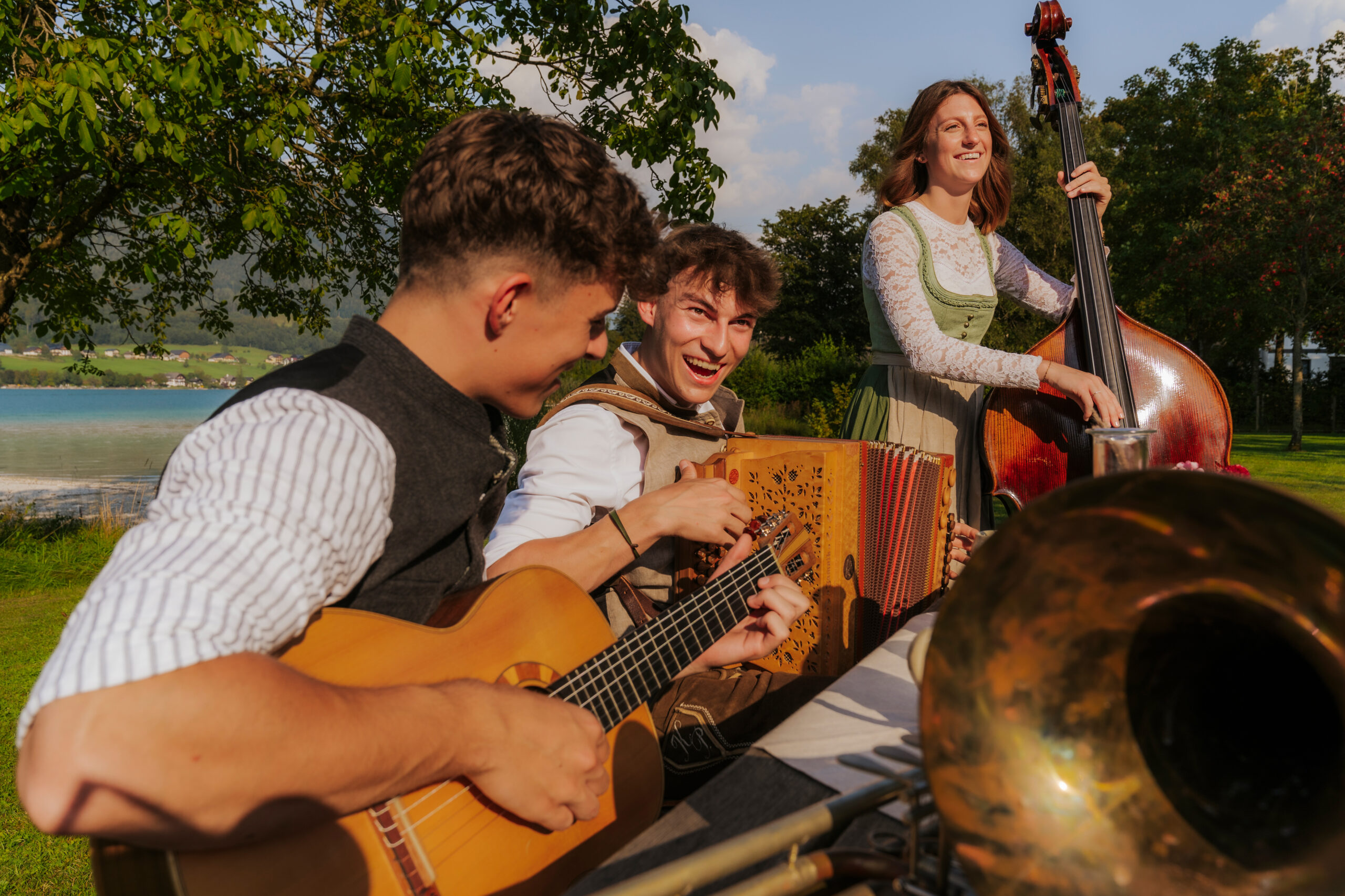 Drei junge Musiker spielen Gitarre, Akkordeon und Kontrabass an einem sonnigen Tag im Freien, lächeln und haben Spaß an der Musik.