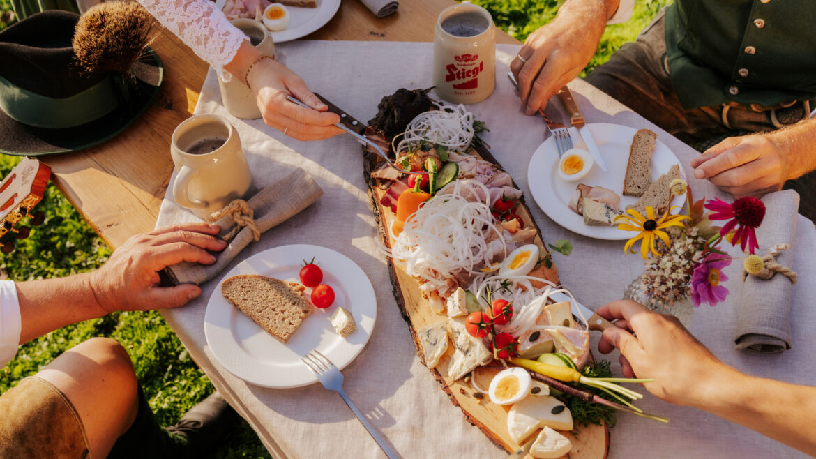 Menschen in traditioneller österreichischer Kleidung teilen sich im Freien eine rustikale Holzplatte mit Fleisch, Eiern, Brot und Gemüse.