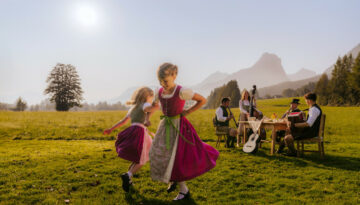 Zwei Mädchen in traditionellen österreichischen Kleidern tanzen im Gras, während Musiker an einem Tisch in einer sonnigen Berglandschaft spielen.
