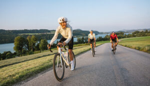 Drei Radfahrer fahren auf einer Landstraße in der Nähe eines Sees, umgeben von grünen Hügeln und Bäumen unter einem klaren Himmel.
