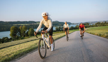 Drei Radfahrer fahren auf einer Landstraße in der Nähe eines Sees, umgeben von grünen Hügeln und Bäumen unter einem klaren Himmel.