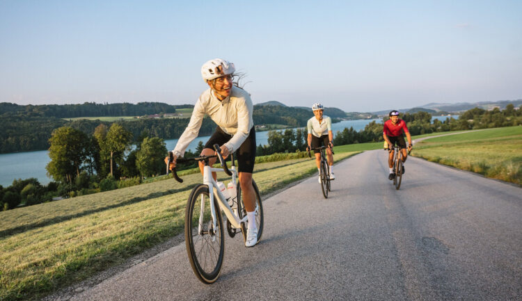 Drei Radfahrer fahren auf einer Landstraße in der Nähe eines Sees, umgeben von grünen Hügeln und Bäumen unter einem klaren Himmel.