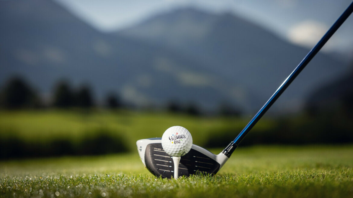 Ein Golfschläger, der so positioniert ist, dass er einen Golfball auf einem grasbewachsenen Platz mit Bergen im Hintergrund schlägt.