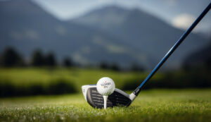 Ein Golfschläger, der so positioniert ist, dass er einen Golfball auf einem grasbewachsenen Platz mit Bergen im Hintergrund schlägt.