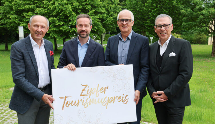 Vier Männer in Anzügen halten ein Schild mit der Aufschrift Zipfer Tourismuspreis im Freien mit grünen Bäumen im Hintergrund.