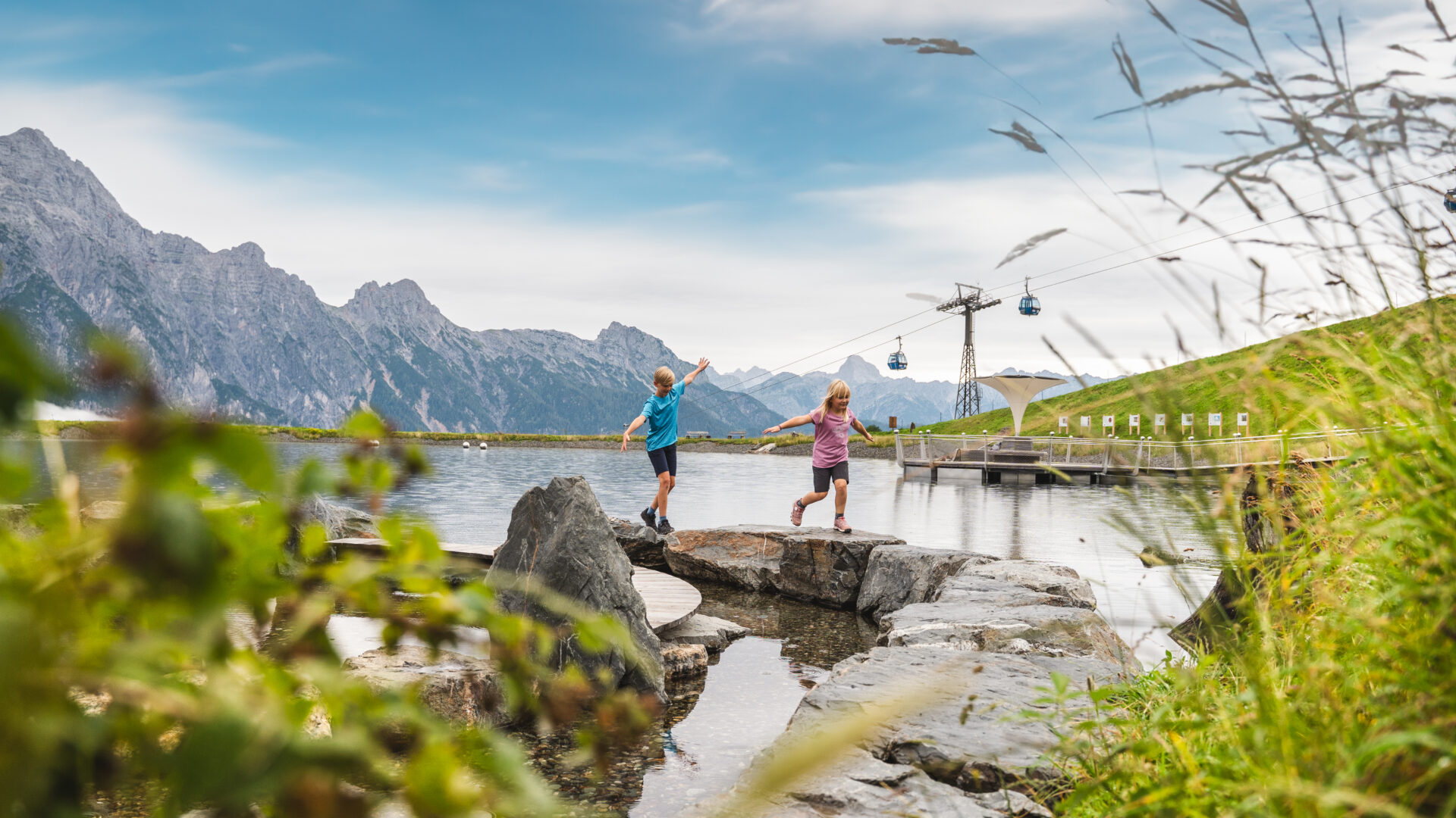 Zwei Kinder balancieren auf Felsen an einem Bergsee, im Hintergrund sind grasbewachsene Hügel und Seilbahnen zu sehen.