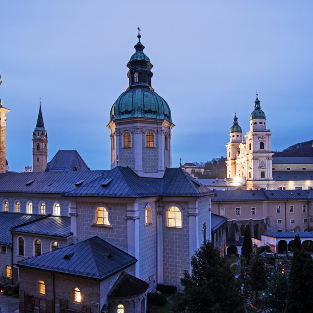 Historische Barockgebäude und Kirchtürme in Salzburg, Österreich, die in der Abenddämmerung vor blauem Himmel beleuchtet sind.