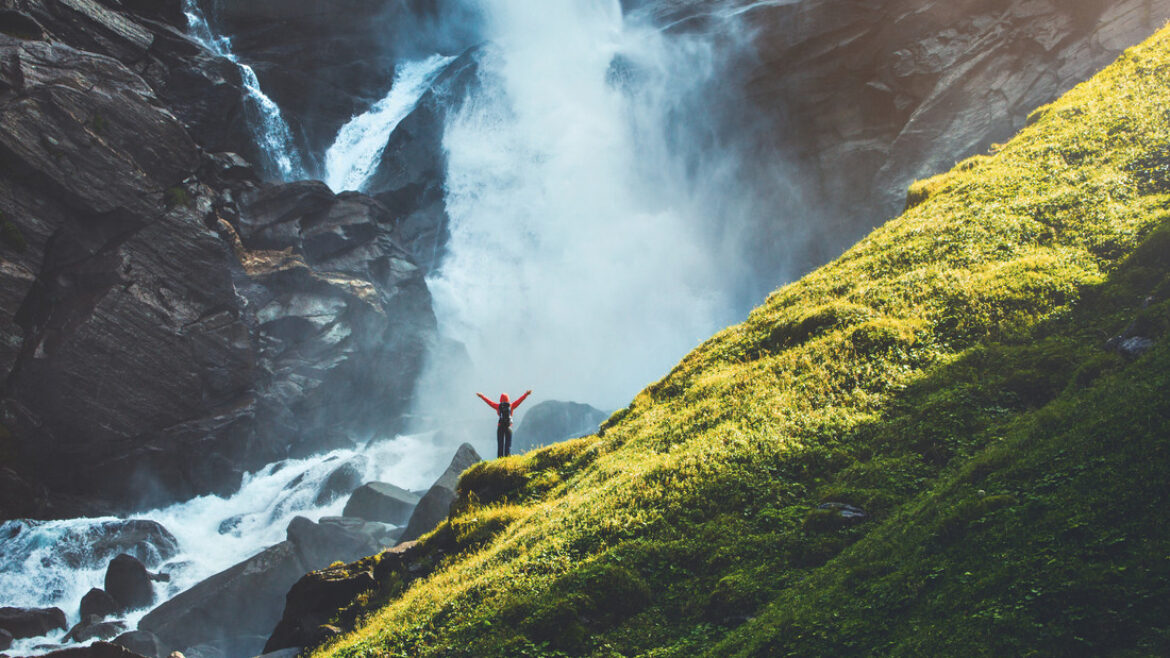 Eine Person mit erhobenen Armen steht auf Felsen in der Nähe eines Wasserfalls, neben einem steilen, sonnenbeschienenen grünen Hang.