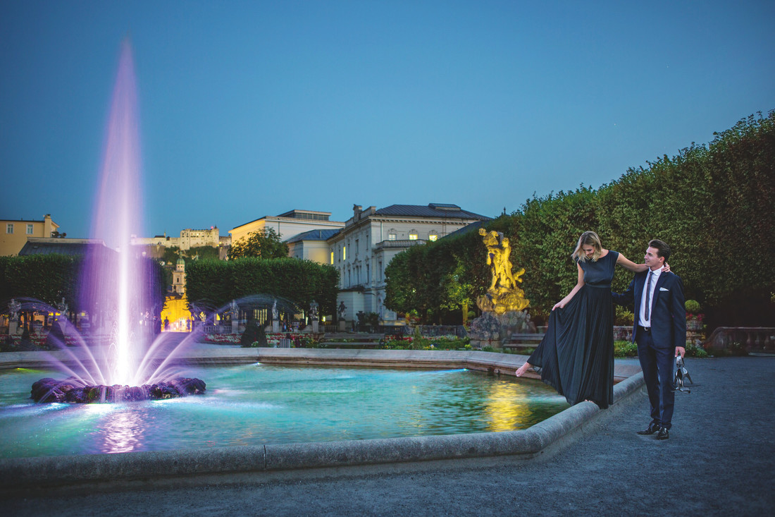 Ein elegant gekleidetes Paar steht in der Abenddämmerung in einem Garten neben einem violett leuchtenden Springbrunnen.