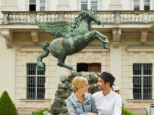 Vor einem prächtigen Gebäude sitzt ein lächelndes Paar vor einer Statue eines geflügelten Pferdes.