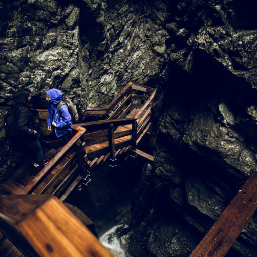 Zwei Menschen in Regenjacken stehen auf einer Holzbrücke in einer felsigen, engen Schlucht.