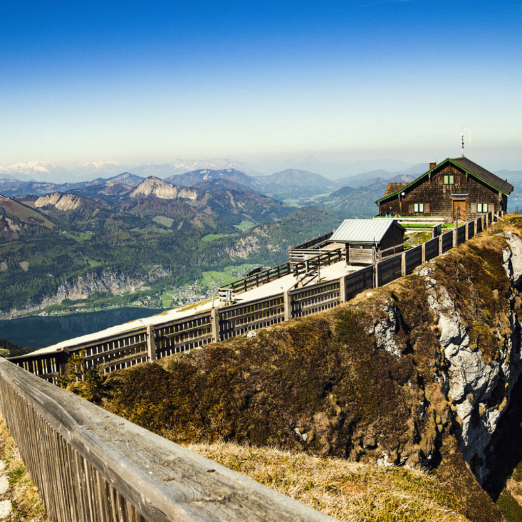 Auf einem Bergfelsen thront eine Holzhütte, von der aus man unter blauem Himmel einen Panoramablick auf Täler und ferne Gipfel hat.