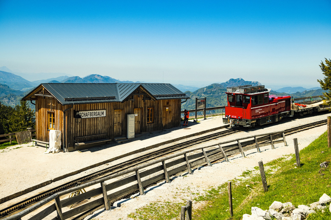 Ein Bahnhof aus Holz mit einem roten Zug, Bergen im Hintergrund und einem strahlend blauen Himmel darüber.