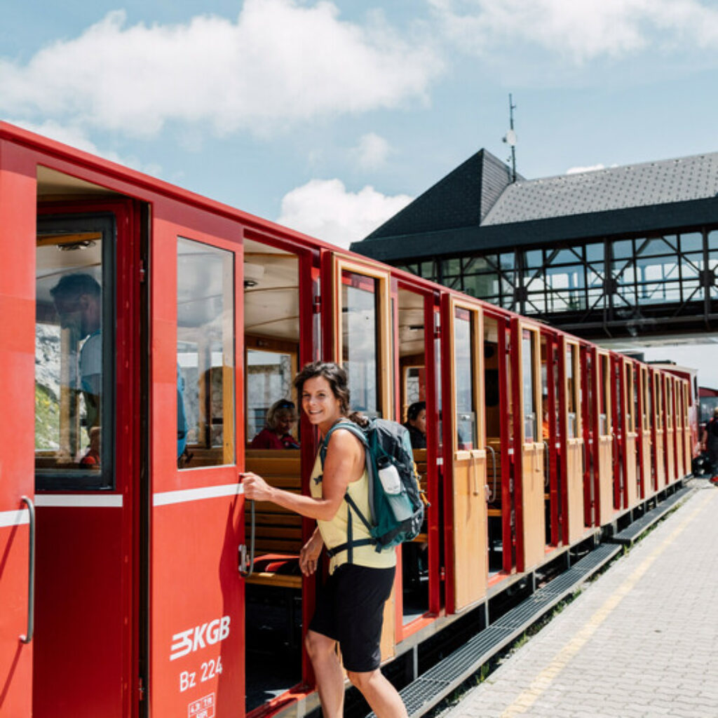 An einem sonnigen Tag steigt eine Frau mit einem Rucksack an einem Bahnhof im Freien in einen roten Zug ein.