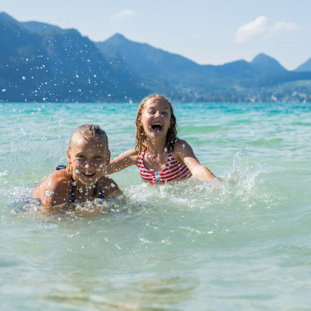 Zwei lächelnde Kinder planschen und spielen im klaren Seewasser, im Hintergrund sind Berge zu sehen.