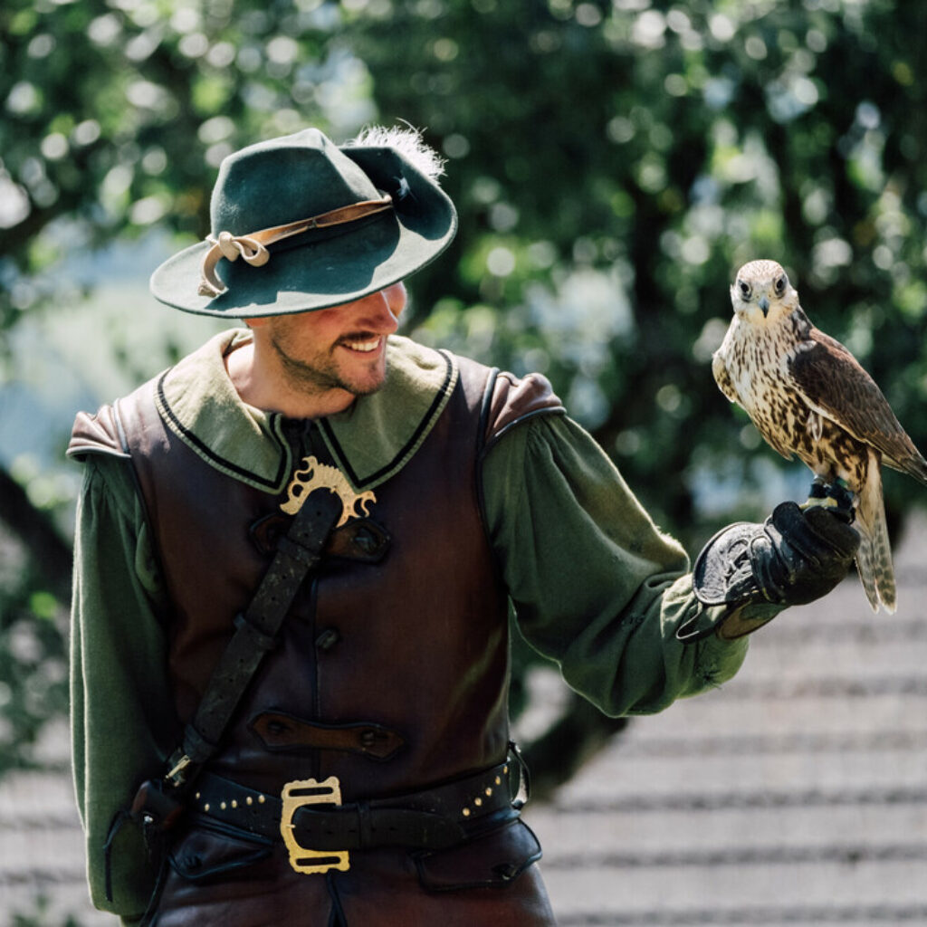 Ein Mann in mittelalterlicher Tracht, der einen Falken auf seiner behandschuhten Hand hält und im Freien vor dem Hintergrund von Bäumen steht.