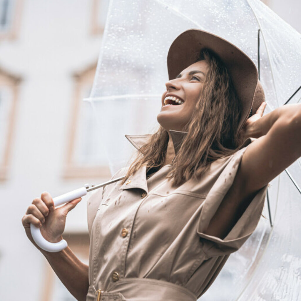 Eine Frau, die im Regen lächelt, einen durchsichtigen Regenschirm in der Hand hält und einen beigen Trenchcoat sowie einen braunen Hut trägt.