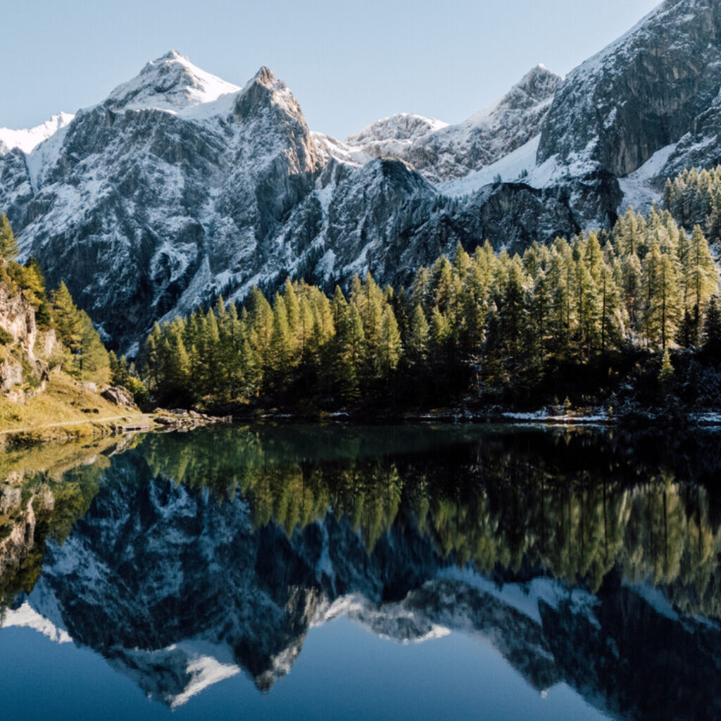 Schneebedeckte Berge und Kiefern, die sich in einem ruhigen See unter einem strahlend blauen Himmel spiegeln.