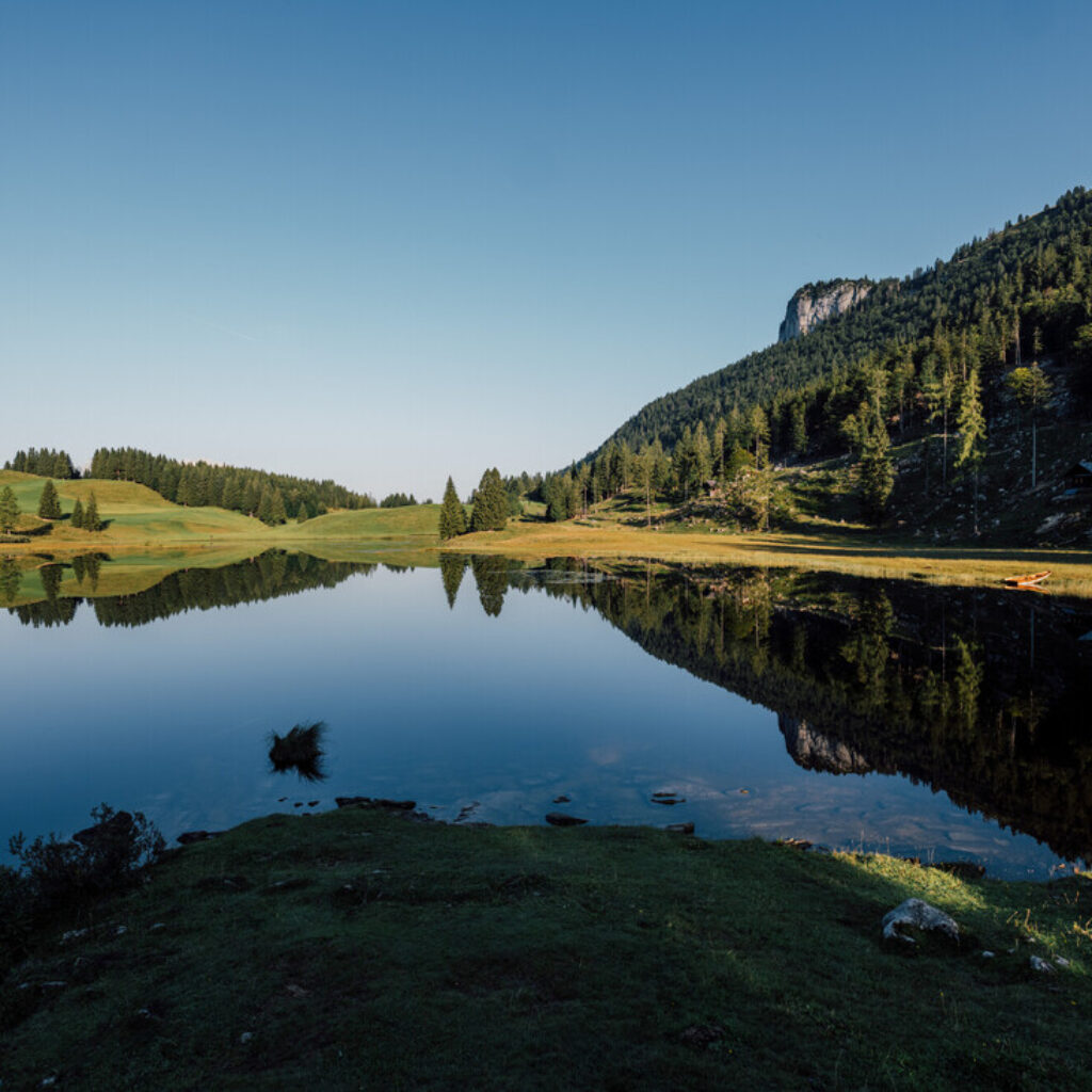 Ein ruhiger See spiegelt grüne Hügel und bewaldete Berge unter einem strahlend blauen Himmel wider.