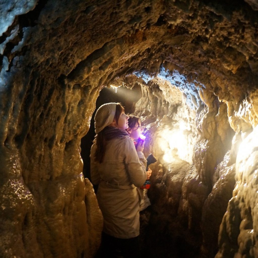 Zwei Menschen erkunden einen schmalen, felsigen Höhlengang, der von Taschenlampen und natürlichen Mineralformationen beleuchtet wird.