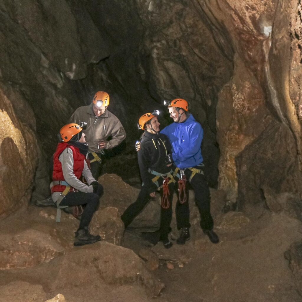 Vier Personen mit Helmen und Stirnlampen unterhalten sich in einer felsigen Höhle.