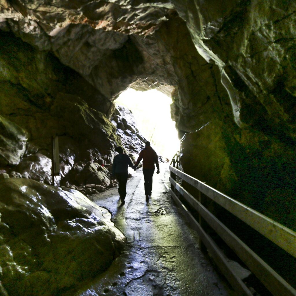 Zwei Menschen, die sich an den Händen halten, gehen auf den hell erleuchteten Eingang eines felsigen Höhlentunnels mit einem Holzgeländer zu.