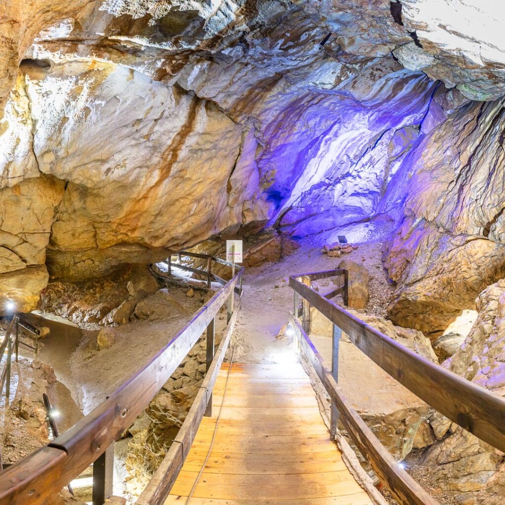 Ein Holzsteg führt durch eine hell erleuchtete Höhle mit felsigen Wänden, und in der Ferne ist eine violette Beleuchtung zu sehen.