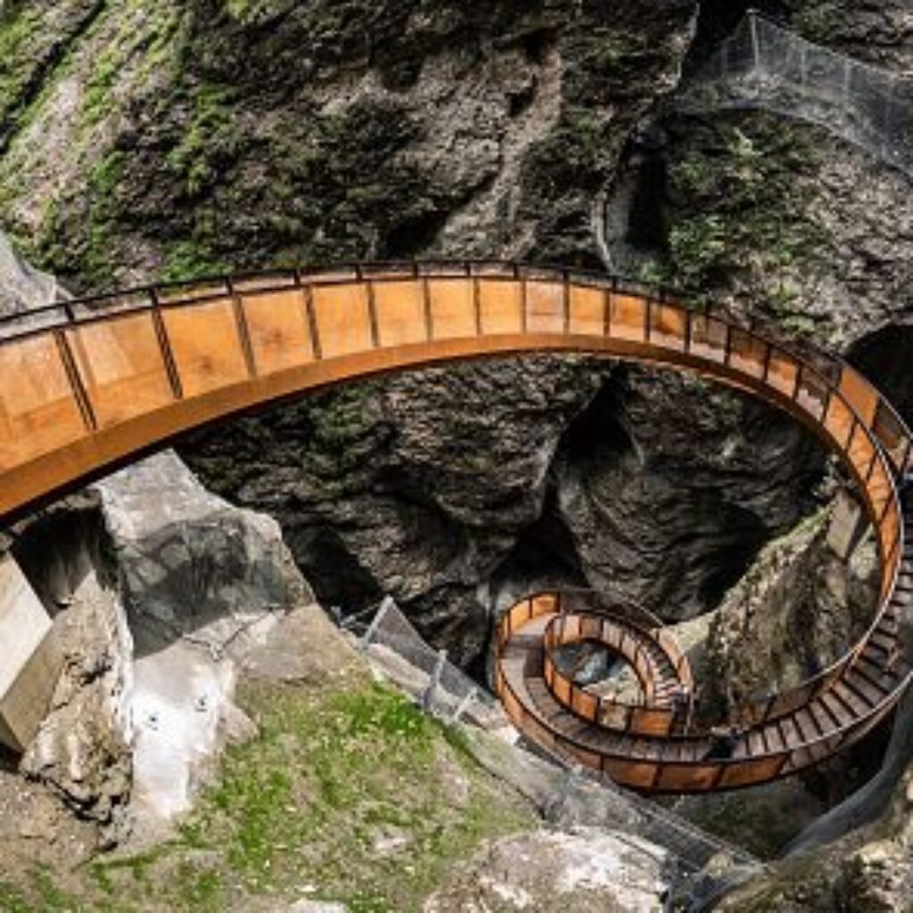 Ein spiralförmiger Holzsteg schlängelt sich entlang einer felsigen Klippe, umgeben von Grün und mit Sicherheitsnetzen gesicherten Absperrungen.