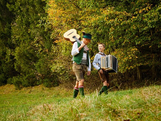 Zwei Menschen in traditioneller österreichischer Tracht gehen draußen spazieren; der eine trägt eine Gitarre, der andere ein Akkordeon.