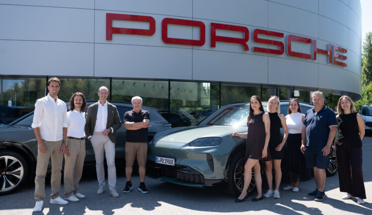 Eine Gruppe von Menschen, die an einem sonnigen Tag vor einem Porsche-Autohaus und einem Porsche-Sportwagen stehen.