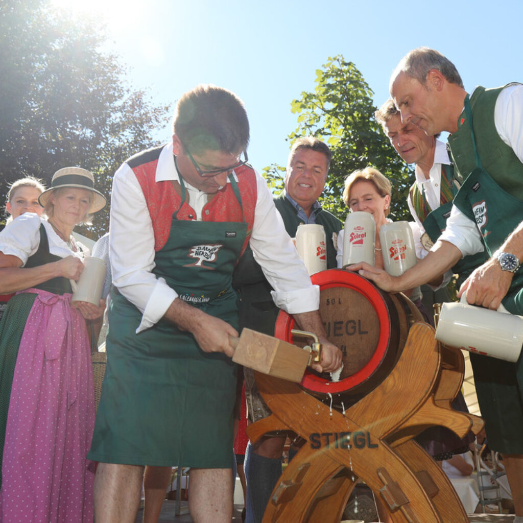 Menschen in österreichischer Tracht zapfen draußen ein Bierfass an, halten Bierkrüge in den Händen und lächeln im Sonnenlicht.