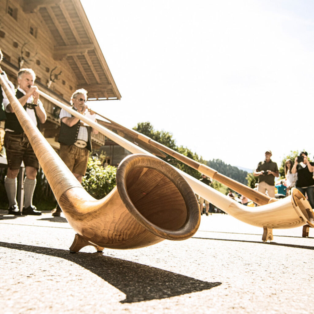 Vier Personen in österreichischer Tracht spielen bei einer Kulturveranstaltung im Freien auf langen hölzernen Alphörnern.