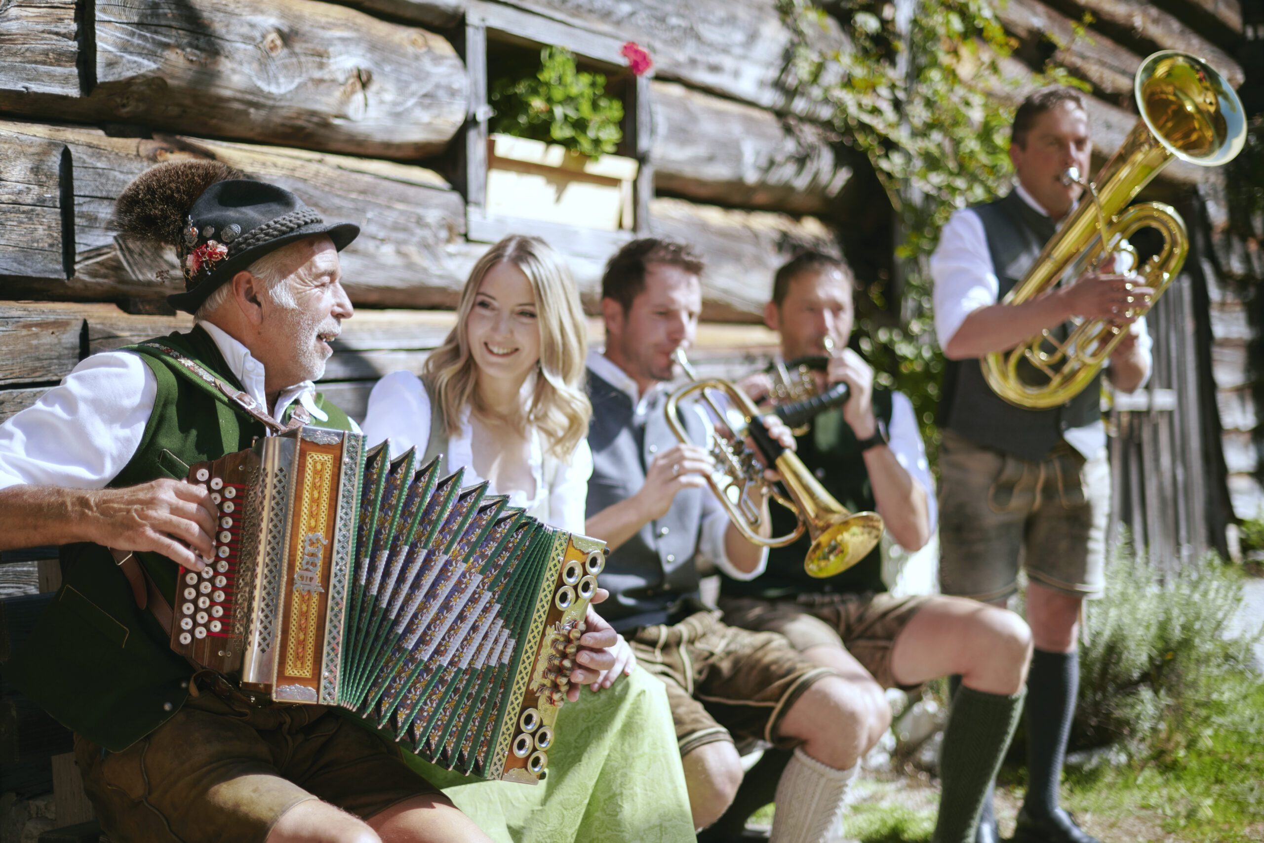Fünf Menschen in traditioneller österreichischer Tracht spielen im Freien neben einem Holzgebäude auf Instrumenten, lächeln und genießen die Musik.