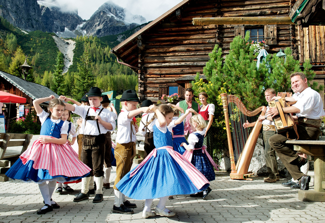 Kinder in traditioneller Tracht tanzen, während Musiker vor einer Holzhütte in den Bergen spielen.