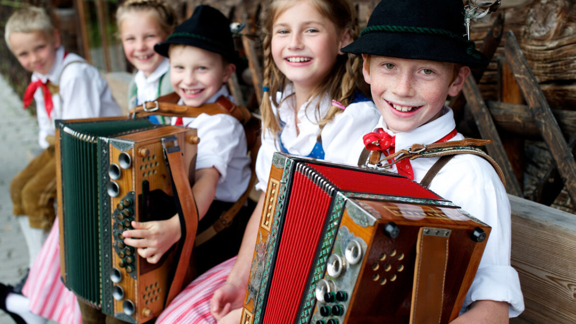 Lächelnde Kinder in traditioneller österreichischer Tracht sitzen auf einer Bank und spielen Akkordeon.
