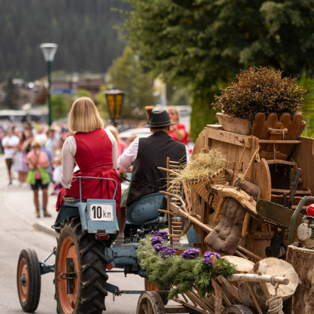 Menschen in traditioneller Tracht fahren bei einer Parade auf einem Traktor und ziehen einen geschmückten Wagen mit rustikalen Gegenständen hinter sich her.