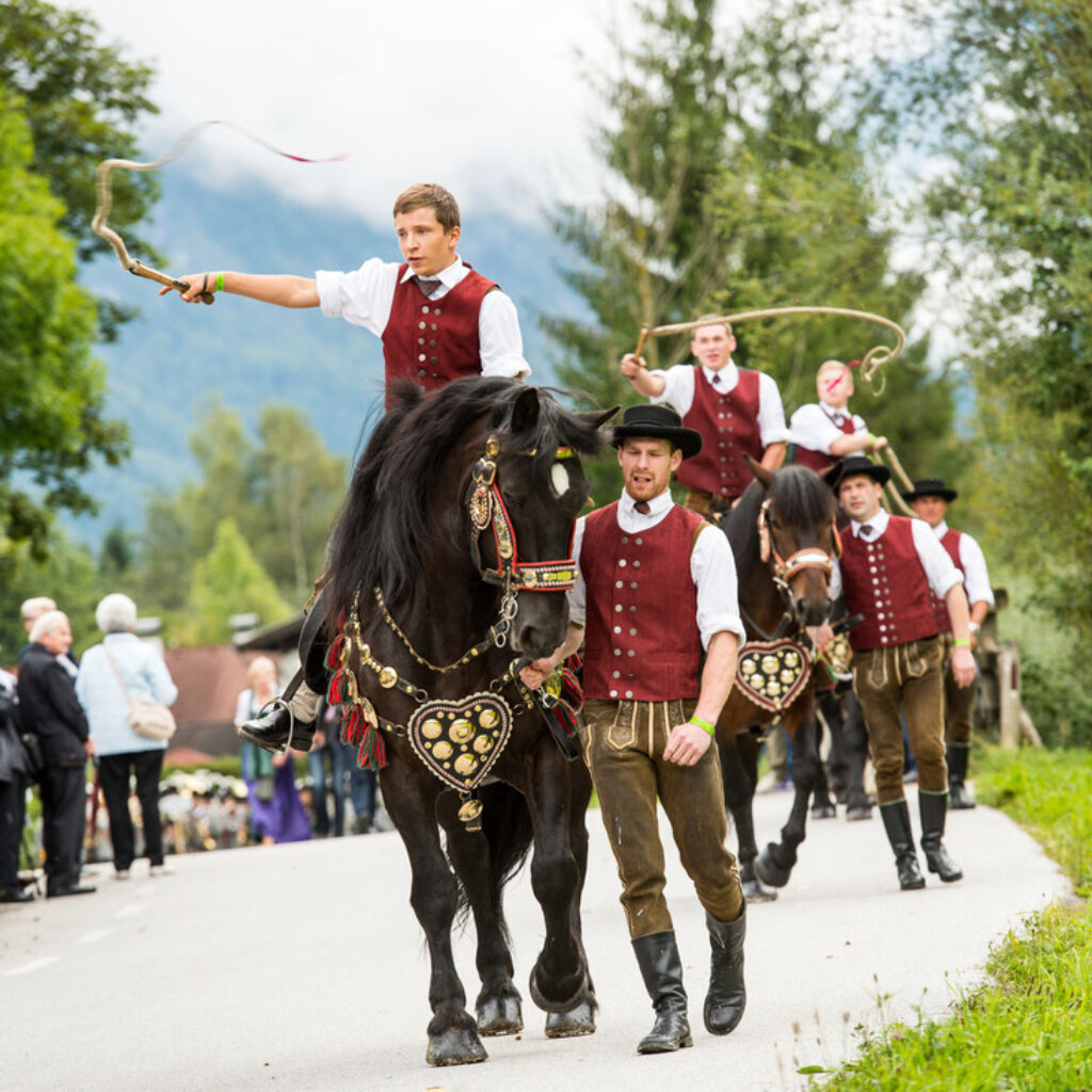 Männer in traditioneller Tracht reiten auf geschmückten Pferden in einer Parade, während die Menschenmassen am Straßenrand zuschauen.