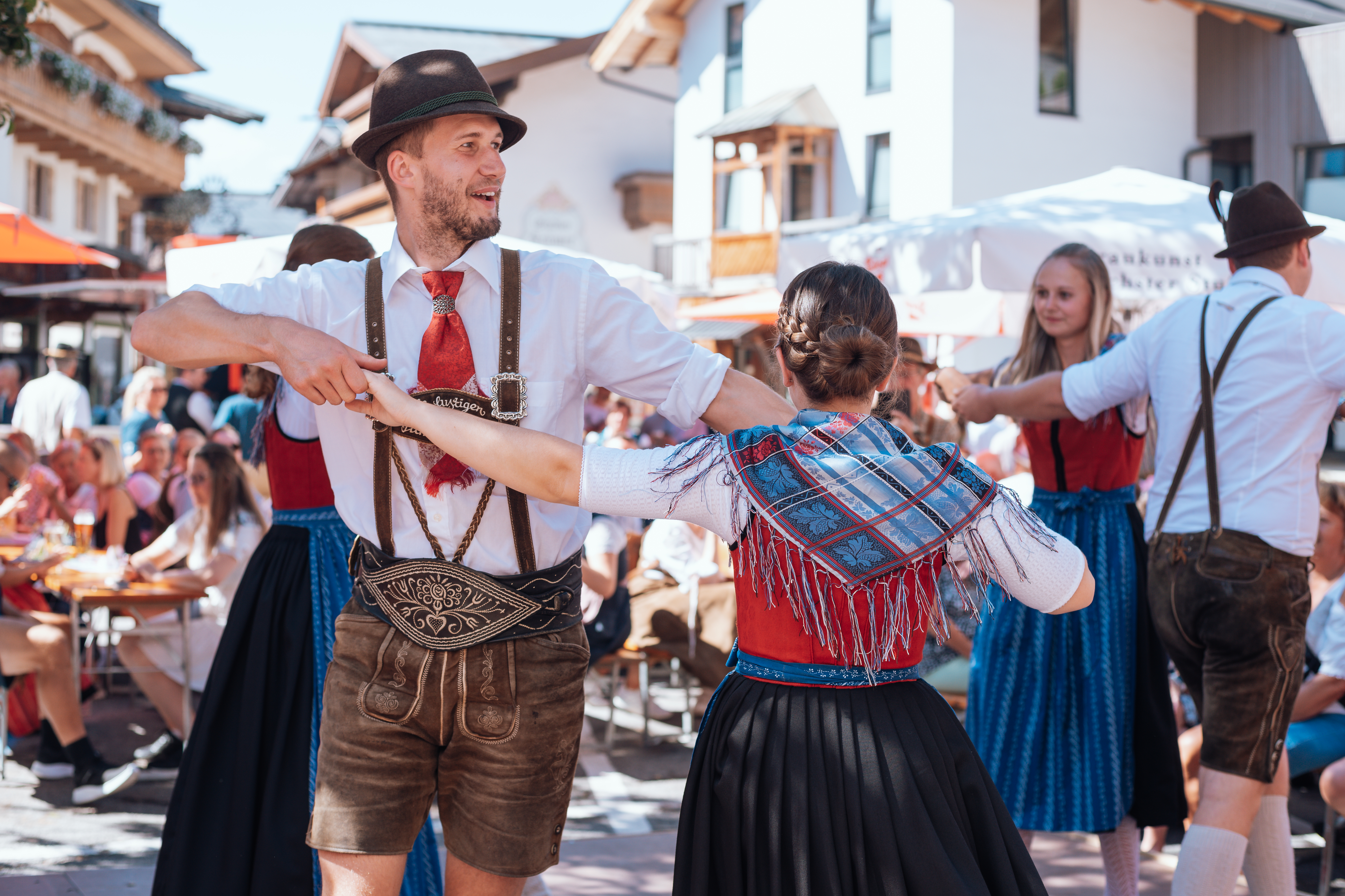 Menschen in traditioneller österreichischer Tracht tanzen bei einer festlichen Veranstaltung im Freien, während Zuschauer in der Nähe sitzen.