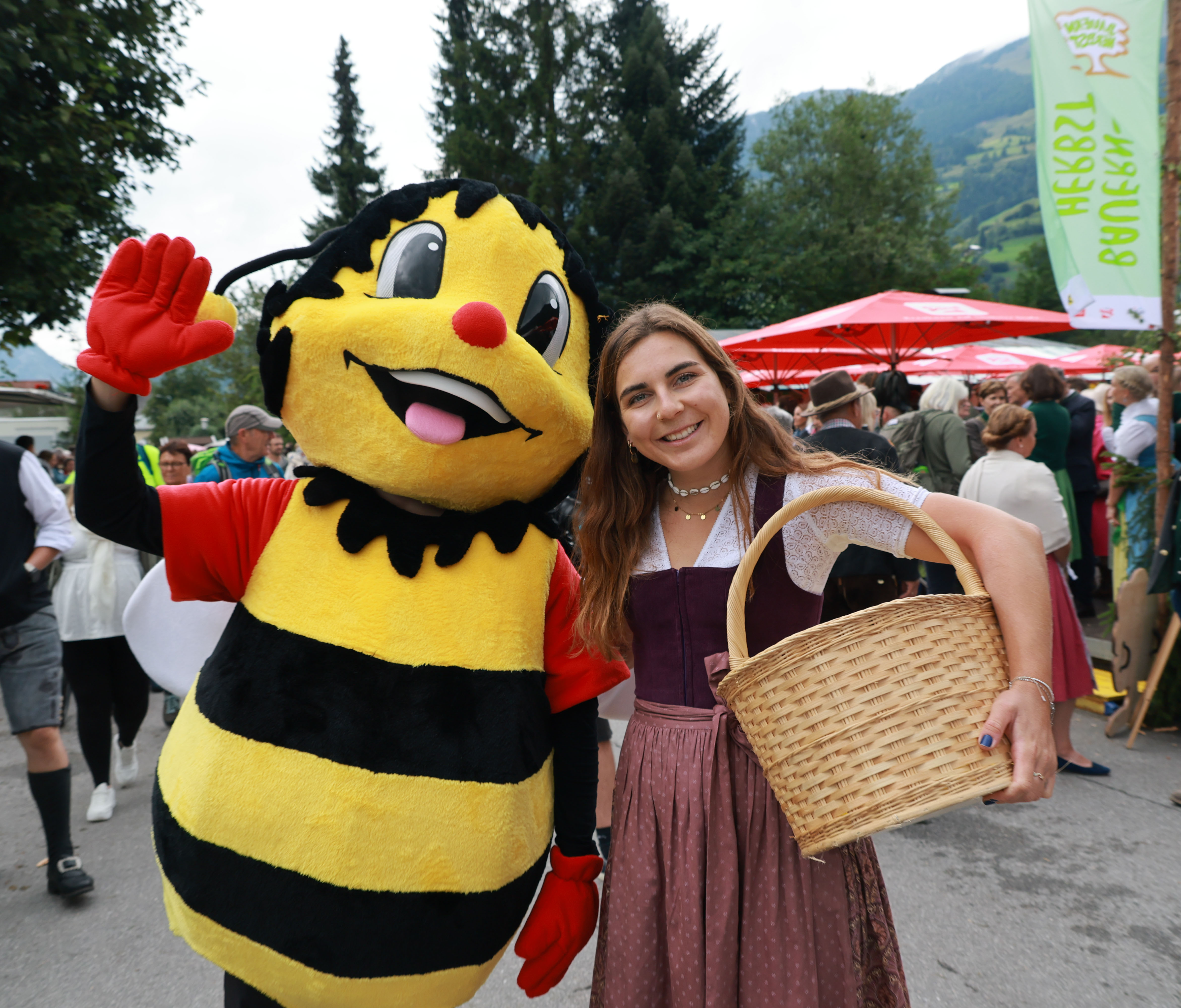 Eine Frau in traditioneller Tracht posiert bei einer festlichen Veranstaltung im Freien mit einem lächelnden Bienen-Maskottchen und hält dabei einen Korb in der Hand.