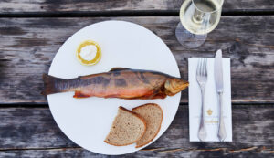 Ein ganzer geräucherter Fisch mit zwei Scheiben Brot, Zitrone und einem Glas Weißwein auf einem rustikalen Holztisch.