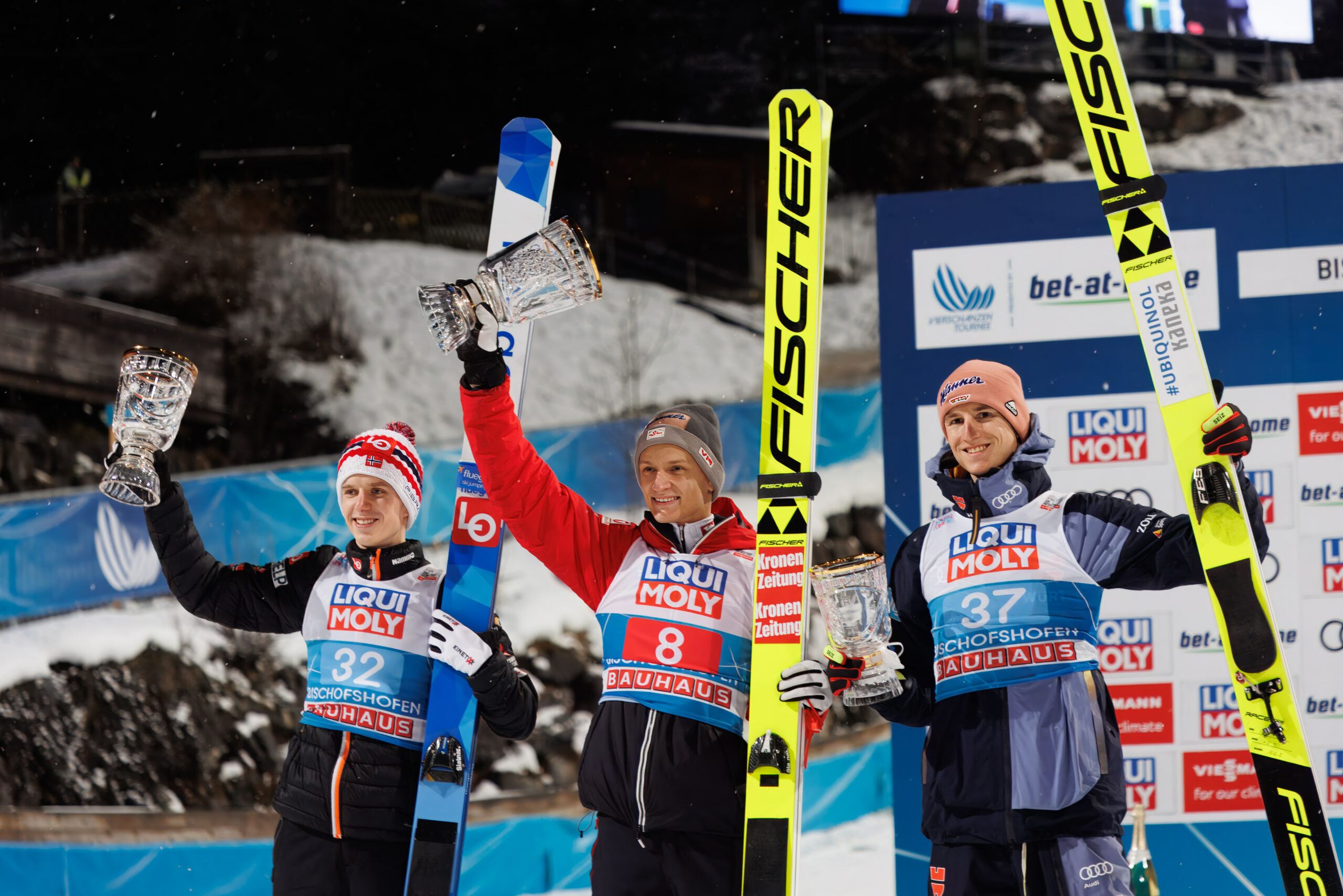 Drei Skispringer stehen nach einem Wettkampf mit Pokalen und Skiern auf einem Podium.