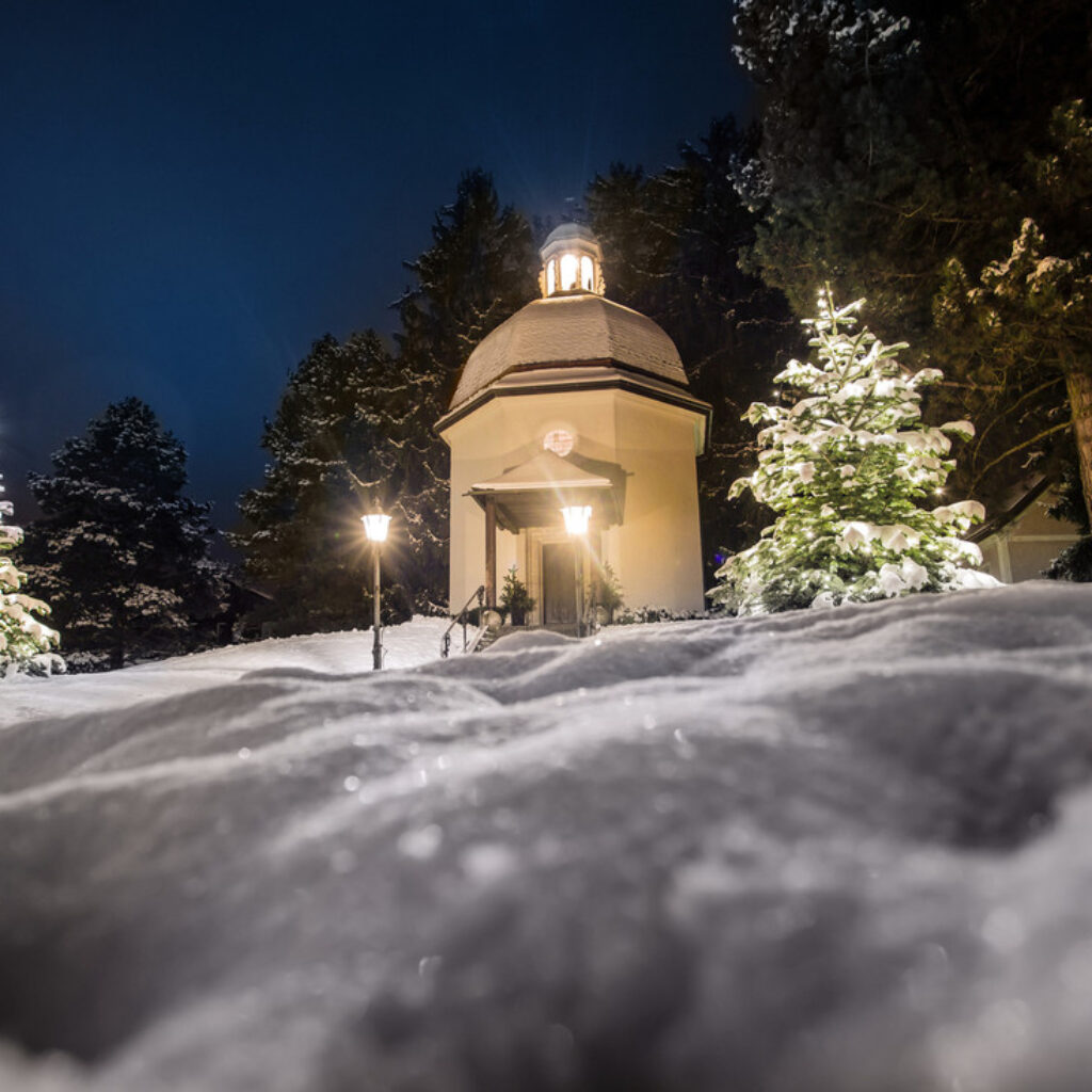 Eine kleine österreichische Kapelle mit Kuppel bei Nacht, umgeben von schneebedeckten Bäumen und leuchtenden Laternen.