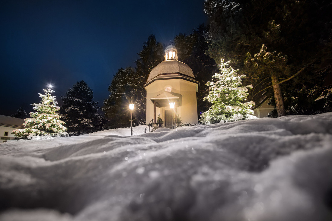 Eine kleine österreichische Kapelle mit Kuppel bei Nacht, umgeben von schneebedeckten Bäumen und leuchtenden Laternen.