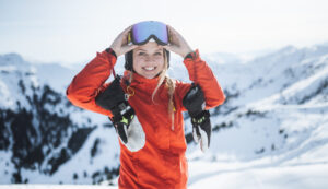 Ein lächelnder Skifahrer in einer roten Jacke und mit einer Skibrille, der auf einem verschneiten Berg steht.