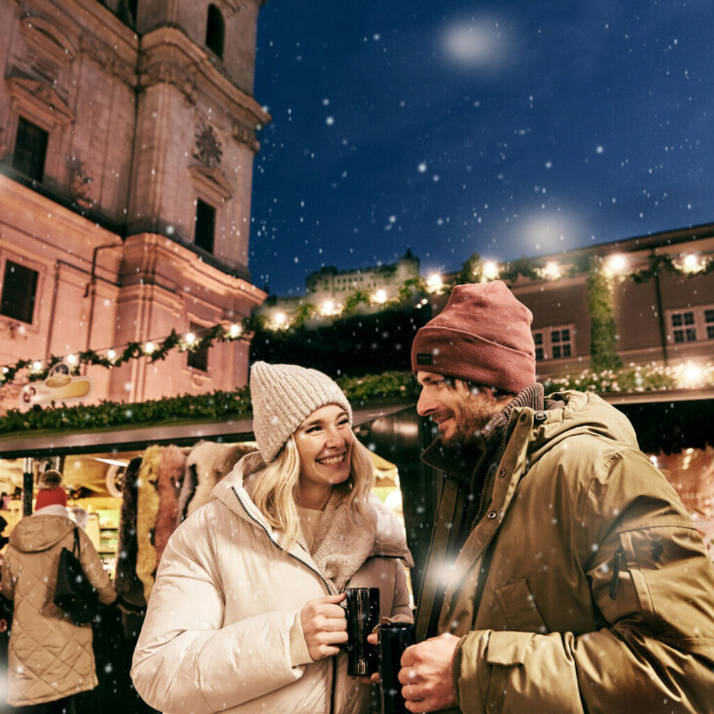 Ein Paar, das auf einem verschneiten österreichischen Weihnachtsmarkt vor einem historischen Gebäude aus Bechern trinkt.