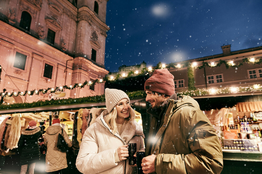 Ein Paar, das auf einem verschneiten österreichischen Weihnachtsmarkt vor einem historischen Gebäude aus Bechern trinkt.