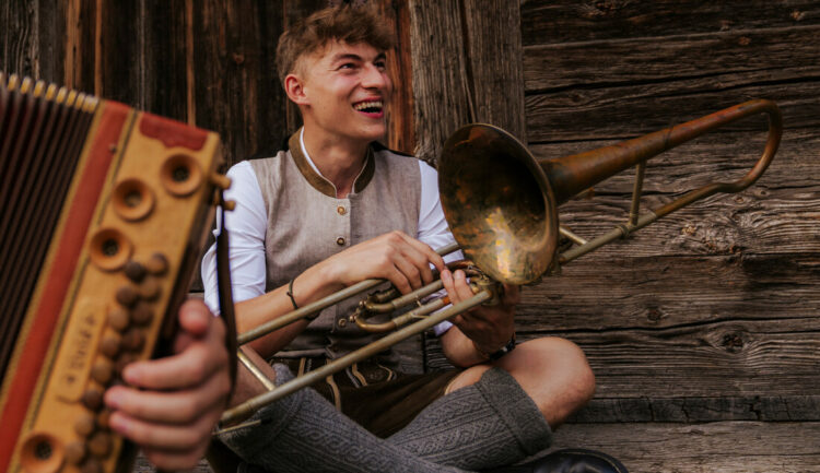 Ein lächelnder Mann in österreichischer Tracht hält eine Posaune in der Hand, im Vordergrund ist teilweise ein Akkordeon zu sehen.