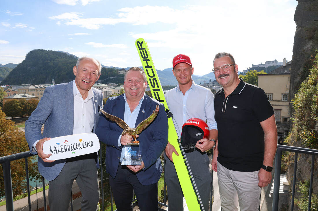 Vier Männer auf einem Balkon mit Blick auf eine österreichische Stadt, mit einer Goldadler-Trophäe, einem Schild und Skisprungausrüstung.