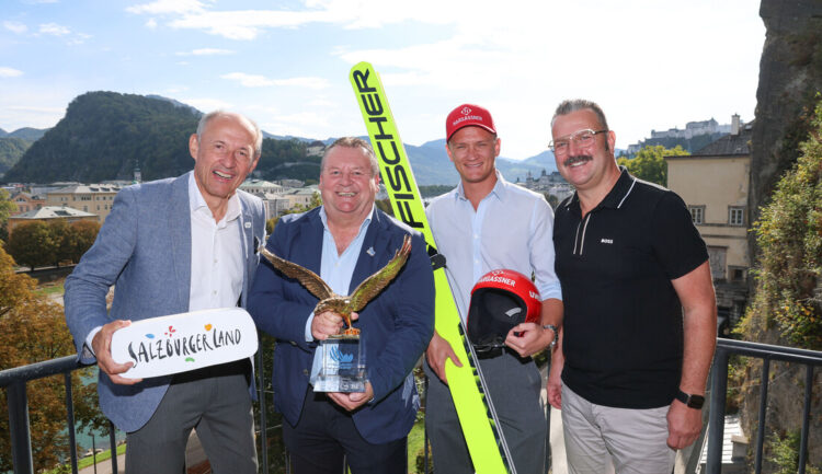 Vier Männer posieren auf einer Terrasse in Salzburg; einer von ihnen hält einen Pokal und ein Paar Skisprungski in der Hand.